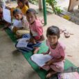 Indian youths wearing pink shirts and white pants seated on green mats with reading materials in hand look up at a photographer snapping their picture.