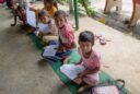Indian youths wearing pink shirts and white pants seated on green mats with reading materials in hand look up at a photographer snapping their picture.