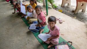 Indian youths wearing pink shirts and white pants seated on green mats with reading materials in hand look up at a photographer snapping their picture.
