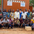 A group of Togan youths and middle-aged men pose with Salesian Missionaries in front of a ochre-colored building with greenery in the background.