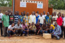 A group of Togan youths and middle-aged men pose with Salesian Missionaries in front of a ochre-colored building with greenery in the background.