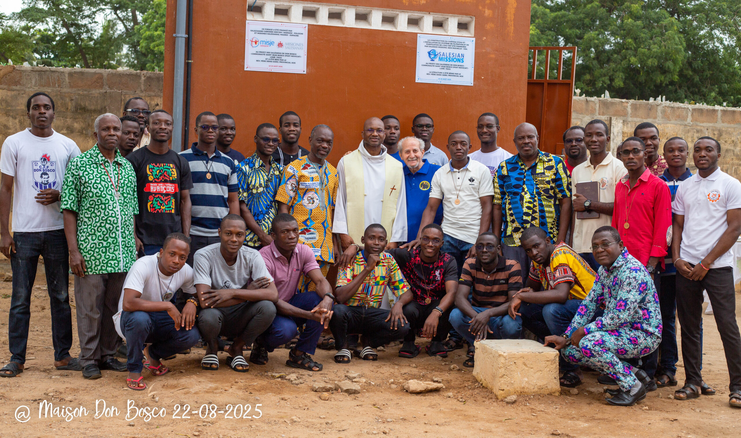 A group of Togan youths and middle-aged men pose with Salesian Missionaries in front of a ochre-colored building with greenery in the background.