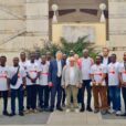 A mixed group of young Ghanaian men and middle-aged Italian businessmen pose for a group portrait in front of a large limestone building.