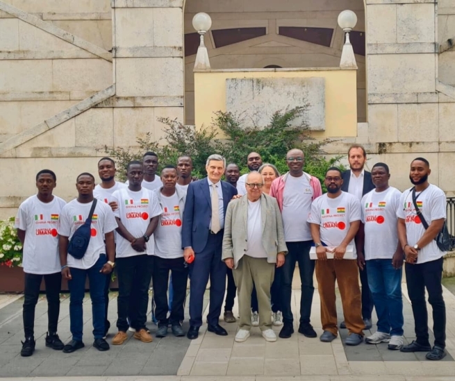 A mixed group of young Ghanaian men and middle-aged Italian businessmen pose for a group portrait in front of a large limestone building.
