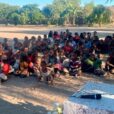 A group photo of dozens of youth seated on a dirt gathering area under the shade of trees.