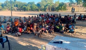 A group photo of dozens of youth seated on a dirt gathering area under the shade of trees.