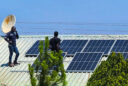 Two Zambian men standing on a metal-roofed building in full sun while installing solar panels. There are green evergreen trees in the foreground.